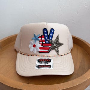 May include: A beige trucker hat with a peace sign, stars, and a flower. The peace sign is red, white, and blue. The hat has a mesh back and a decorative cord along the brim. The hat has a label that says "World Famous Foam Front Trucker".