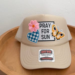 Op de afbeelding: Beige truckerpet met een mesh achterkant en een gebogen klep. De voorkant heeft de tekst "PRAY FOR SUN", een roze bloem, een blauw-wit hart en een oranje vlinder. Voorzien van een drukknoopsluiting.