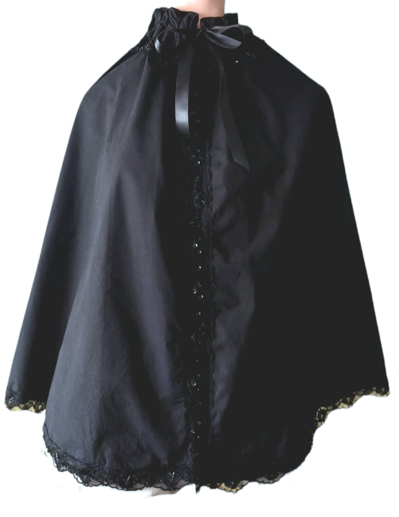 May include: A black cape with a lace trim and a black satin ribbon tied at the neck. The cape has a high collar and is made of a soft, flowing fabric.