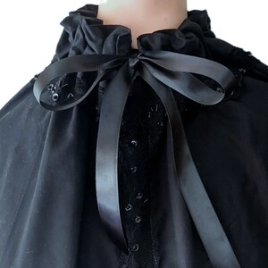May include: A black velvet cape with a ruffled collar and a black satin ribbon tied at the neck. The cape is decorated with black sequins.