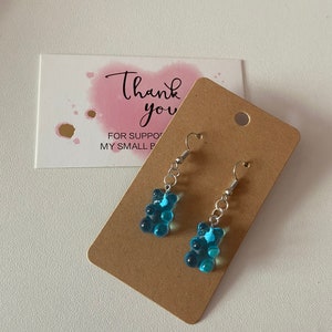 May include: A pair of blue gummy bear earrings with silver hooks. The earrings are displayed on a brown card with the text "Thank you" and "For supporting my small business".