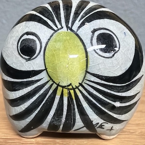 May include: A ceramic owl figurine with a cracked, off-white glaze. The owl features black and white striped wings, large black eyes, and a yellow oval face. The base has three small feet.