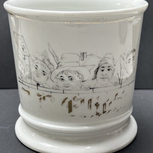 May include: White ceramic mug with a wide base and a slightly flared rim. The mug features a black and white illustration of several faces peering over a fence, with text in gold lettering below the illustration. The mug is a vintage style.