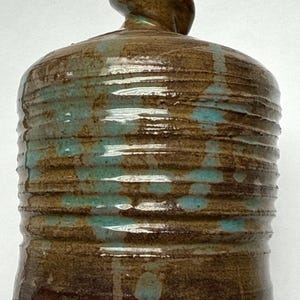 May include: A handmade ceramic vase with a brown and teal glaze. The vase has a rounded body with horizontal ridges and a small, rounded top. Teal glaze drips down the sides, creating a unique, textured appearance. The vase is a decorative item.
