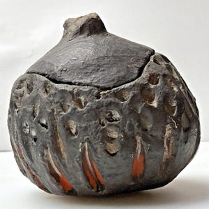 May include: A dark gray, handcrafted ceramic jar with a lid. The jar has a textured surface with carved patterns and red accents. The lid has a unique, irregular shape. The jar is likely used for storage or decorative purposes.