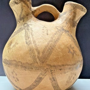May include: A tan-colored, double-spouted ceramic vessel with a handle. The vessel features dark brown geometric patterns and decorative dots. The rim and handle show signs of wear, suggesting age. The overall design is simple and rustic.