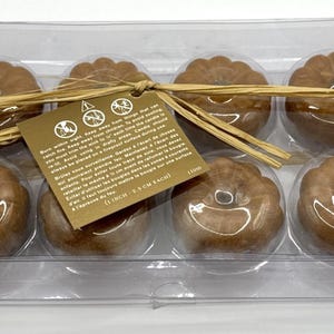 May include: A clear plastic box filled with eight brown pumpkin-shaped candles. The candles are arranged in two rows of four. The box is tied with a brown ribbon and has a gold label with safety instructions.
