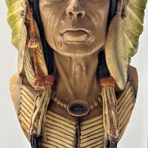 May include: A detailed bust of a Native American chief, featuring a headdress with feathers and intricate beadwork. The sculpture is painted in earthy tones with a green base. The figure has a serene expression and a beaded necklace.