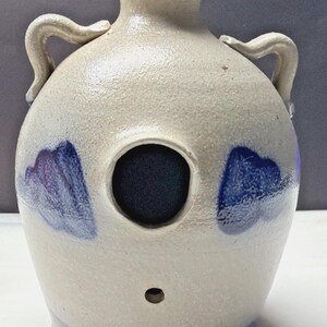 May include: A cream-colored ceramic jug with two side handles, a narrow neck, and a wide body. The jug features a large circular opening in the center, a smaller hole below it, and blue painted accents on the sides and base with a glossy finish.