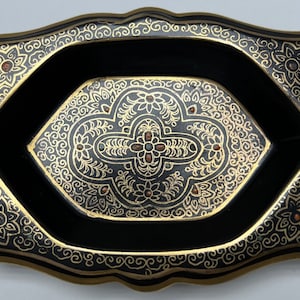Op de afbeelding: Een rechthoekige zwarte tray met een sierlijk gouden bloemenontwerp. De tray heeft een ovale centrale vorm met een gouden bloemenpatroon en is versierd met kleine rode en groene juwelen. De randen zijn ook versierd met gouden krullen.