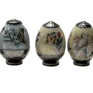 May include: Five decorative egg-shaped containers with silver-colored bases and lids. Each egg features a different nature-themed design, including a swan, butterflies, and a bird. The eggs are painted in pastel colors.