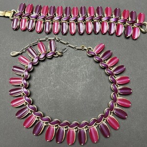 May include: A set of vintage jewelry featuring a necklace and bracelet. Both pieces are made of gold-toned metal and feature a repeating pattern of oval-shaped pink and purple stones.