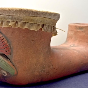 May include: A terracotta-coloured, foot-shaped vessel with a flared opening and a smaller, cylindrical opening. The vessel features a decorative head with a headdress and a fan-shaped element. The opening is adorned with a tan, fringed material.