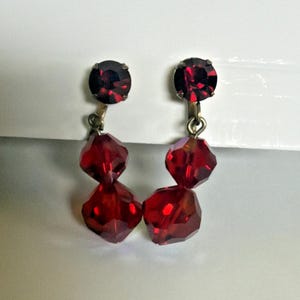 May include: A pair of vintage clip-on earrings featuring a dark red, faceted, teardrop-shaped bead design. Each earring has a small, round, dark red stone at the top. The earrings are set against a white background.
