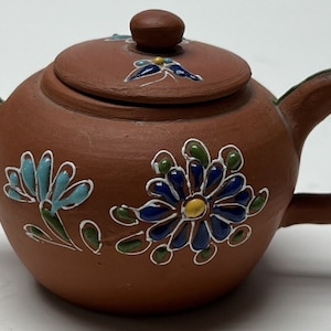 May include: A small terracotta teapot with a lid, decorated with hand-painted blue, green, and yellow flowers.