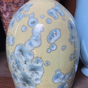 Puede incluir: Un jarrón de cerámica amarilla con patrones cristalinos azules. El jarrón tiene una forma redondeada con un cuello estrecho. Los patrones azules se agrupan en varios tamaños sobre la superficie amarilla, creando un efecto decorativo único.