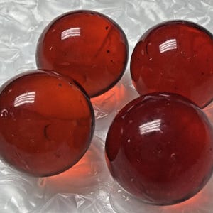 May include: Four translucent, round, red glass spheres. The spheres have a glossy finish and reflect light. The objects are arranged closely together on a crinkled, white surface. The spheres appear to be decorative or collectible items.