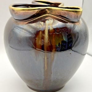 May include: A dark, iridescent ceramic vase with a unique shape. The vase has a wide, rounded body that tapers towards the base. The rim is accented with a gold-colored trim. The surface of the vase reflects light, creating a shimmering effect.