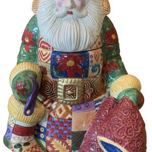 May include: A ceramic Santa Claus figure with a white beard, rosy cheeks, and a burgundy hat. The figure wears a green coat with red floral accents and a patchwork design on the body. Santa holds a sack and a stocking.