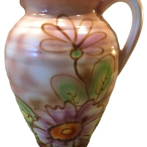 May include: A ceramic pitcher with a floral design. The pitcher is decorated with pink, purple, green, and brown flowers. The pitcher has a handle and a rounded body.