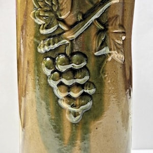 May include: A cylindrical, handmade ceramic vase with a tan and green glaze. The vase features a raised relief design of grapes and leaves. The design is highlighted with white accents. The vase is suitable for home decor.