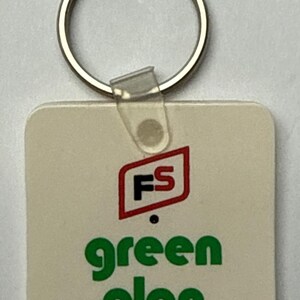 May include: A square, off-white keychain with a silver ring. The keychain features the text "green plan" in green, and a red and black logo that reads "FS".