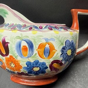 May include: A white ceramic creamer with a red handle and a floral design in shades of blue, orange, yellow, green, and purple.