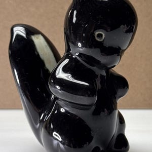 May include: A glossy black ceramic skunk figurine with a white stripe on its tail. The skunk is in a seated position, with its head slightly bowed. The figurine has a smooth, reflective surface.