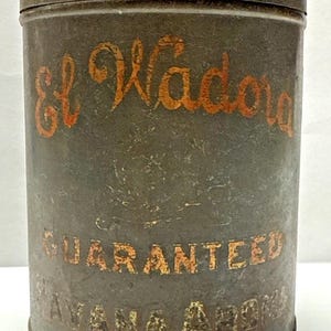 May include: Vintage metal tin with the words "El Wadora" in orange script. The tin is cylindrical with a lid and features the words "GUARANTEED HAVANA AROMA" in orange block letters. The tin has a distressed, aged appearance.