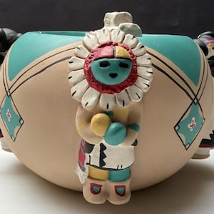 May include: A ceramic pot with a turquoise rim and tan body, adorned with Native American figures. The pot features a central figure with a turquoise face, red and white headdress, and a yellow and blue garment. Additional figures are on the sides.