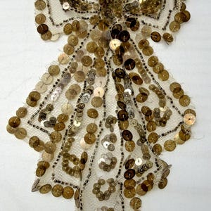 May include: A vintage-style, bow-shaped embellishment. The bow is made of a cream-colored, open-weave fabric and is heavily adorned with gold and silver sequins. The sequins are sewn onto the fabric, creating a shimmering effect. The bow is likely for clothing or accessories.