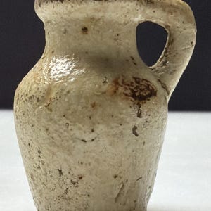 May include: A small, off-white ceramic jug with a handle. The jug has a rounded body and a narrow neck, with a dark brown rim and spots. The handle is a simple loop. The jug appears to be aged or antique.