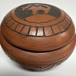 May include: A round, brown, lidded box with a carved design. The lid features a black silhouette of an animal on a hill, surrounded by a rectangular pattern. The box's base has a geometric design. The box is made of a natural material.