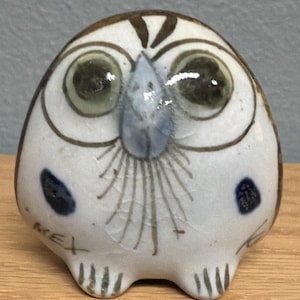 May include: A ceramic owl figurine with a white base, brown accents, and large green eyes. The owl has a blue beak and two blue spots on its sides. The word "MEX" is written on the bottom left side of the owl.