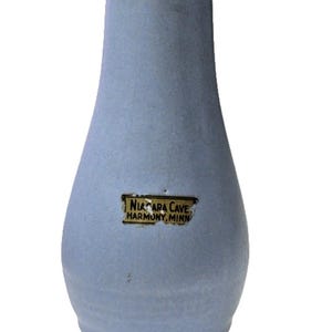 May include: A light blue ceramic vase with a tapered neck and a rounded base. A rectangular sticker is attached to the front, featuring the black text NIAGARA CAVE HARMONY, MINN on a tan background. The vase has a smooth, matte finish throughout.