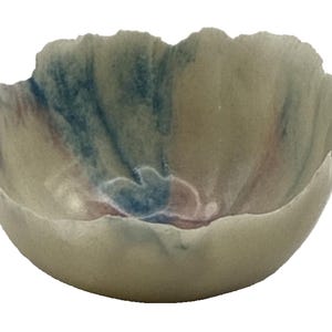 May include: A handmade ceramic bowl with a unique, organic shape. The bowl features a soft, creamy base color with subtle blue, red, and green watercolor-like accents. The rim has a textured, uneven edge, adding to its artistic appeal.