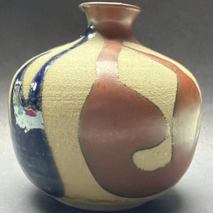 Könnte beinhalten: Kleine, runde Keramikvase mit schmalem Hals. Die Vase hat einen strukturierten, beigefarbenen Hintergrund mit einem Muster aus dunkelroten und blauen geschwungenen Formen. Der obere Teil und der Hals sind dunkelbraun.