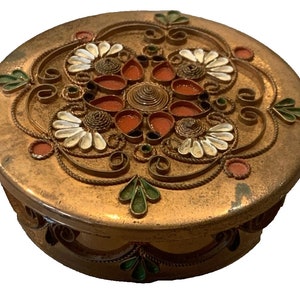 May include: A round, gold-colored trinket box with a decorative design of white, green, and orange paper curls. The box has a lid with a floral pattern.