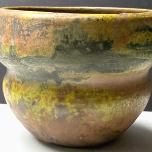 May include: A handcrafted ceramic pot with a wide opening and a bulbous base. The exterior features a mottled, earthy color palette of brown, yellow, green, and orange hues. The pot's surface has a textured appearance, suggesting a rustic, artisanal quality.
