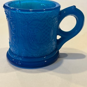 May include: A small, blue glass mug with a decorative embossed pattern. The mug has a single handle and a rounded base.