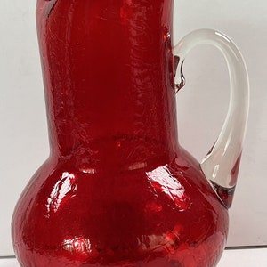 May include: A red glass pitcher with a clear glass handle. The pitcher has a textured, crackled surface.
