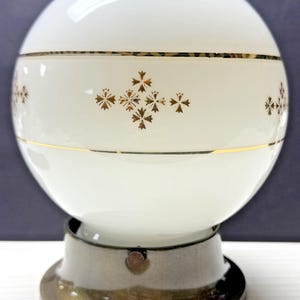 Może przedstawiać: Wintage biała szklana lampa kulista ze złotymi akcentami i kwiatowym wzorem. Lampa stoi na podstawie w kolorze mosiądzu, prezentując klasyczny design. Ta dekoracyjna oprawa oświetleniowa jest idealna do dekoracji wnętrz.