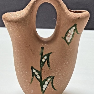 Puede incluir: Un pequeño recipiente de terracota texturizada con un asa central arqueada y dos aberturas en la parte superior. La superficie exterior es de color arcilla marrón claro, decorada con tallos verdes y detalles florales blancos pintados a mano.