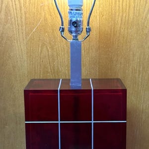 May include: A modern table lamp with a red, cube-shaped base, accented with white lines. The lamp has a silver metal stem and a bright, round light bulb. The lamp is on a silver base.