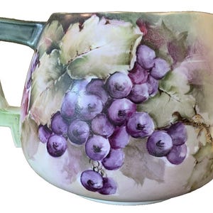 May include: A white porcelain pitcher with a green handle and a hand-painted design of purple grapes and green leaves.