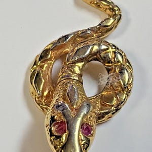 May include: A gold-toned metal snake pendant featuring a coiled body with textured scale detailing and two small, round, pink-colored stones set as eyes. The pendant includes a small circular loop at the top for attachment to a chain or cord.