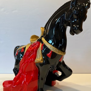 May include: Black ceramic horse figurine with a red and gold saddle and a red cape. The horse is standing on its hind legs with its head tilted down.