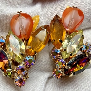 May include: A pair of vintage clip-on earrings featuring a floral design. The earrings showcase a combination of orange, yellow, and iridescent rhinestones. The design includes leaf-shaped and teardrop-shaped stones, with a gold-tone metal setting.