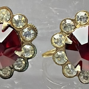 May include: A pair of vintage stud earrings. Each earring features a large, faceted, deep red gemstone at the center, surrounded by a halo of smaller, clear, round gemstones. The settings are gold-toned metal.