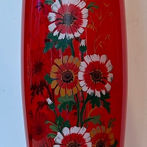 May include: A tall, red glass vase with a gold rim and floral design. The vase features white, red, and yellow flowers with green leaves.
